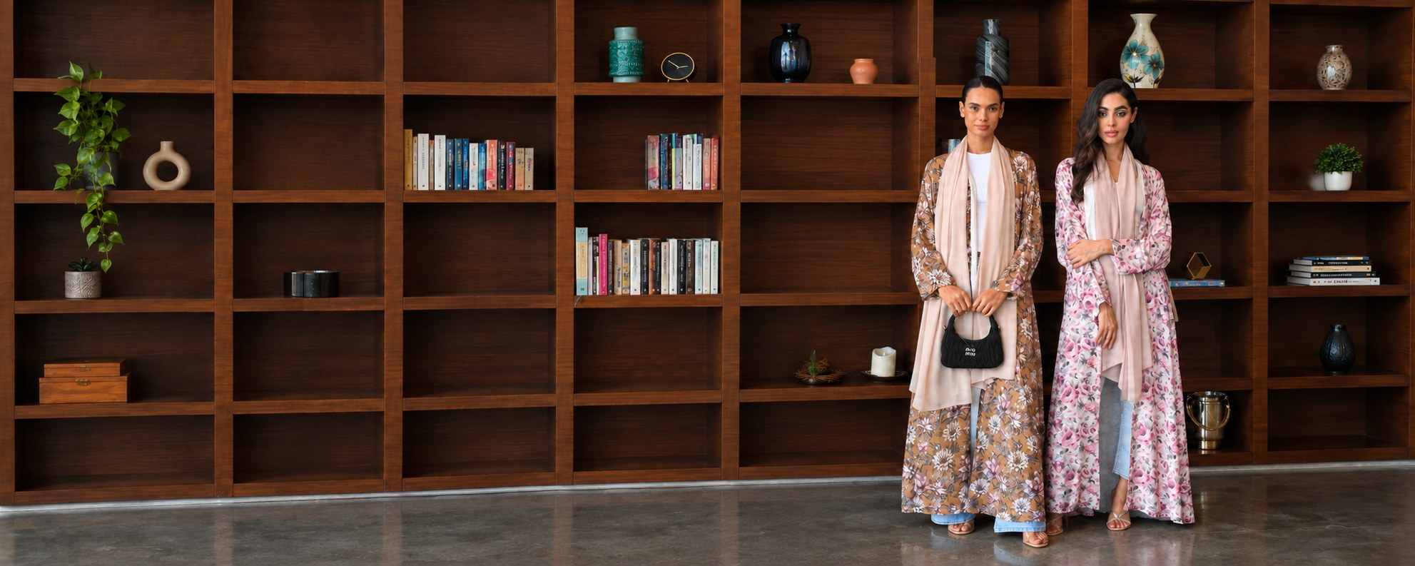 Two women in floral Hiza abayas standing before a wooden bookshelf